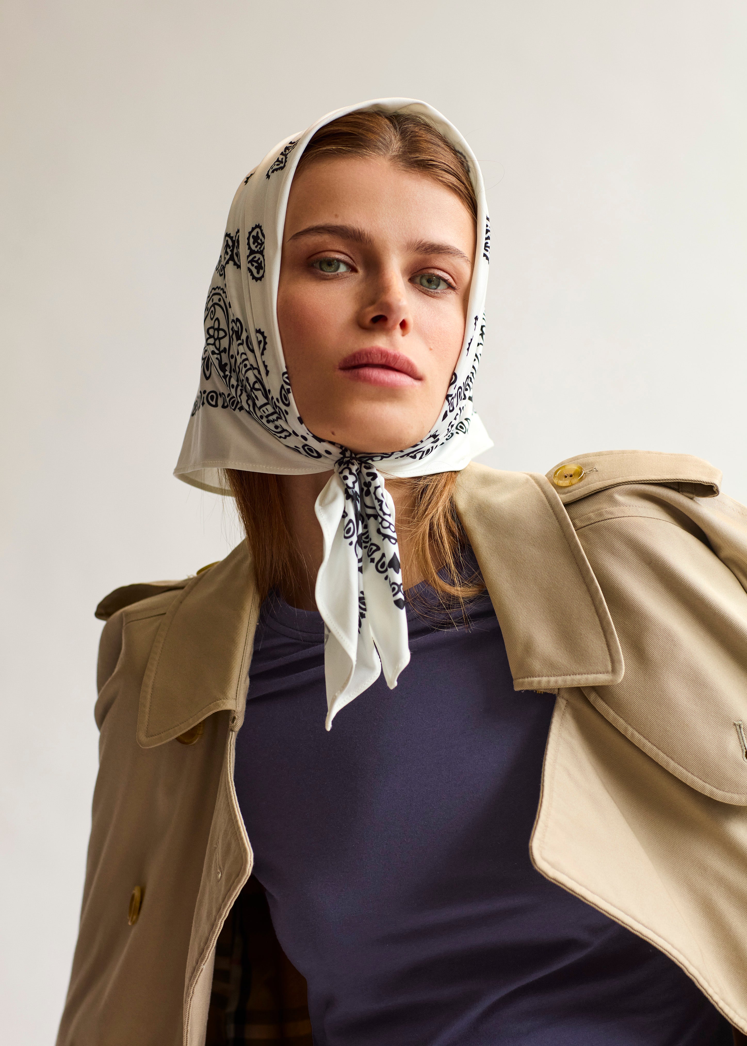 Woman wearing white Lola Swim Bandana UPF 50+ with black paisley print, styled with trench coat for sun-safe elegance.